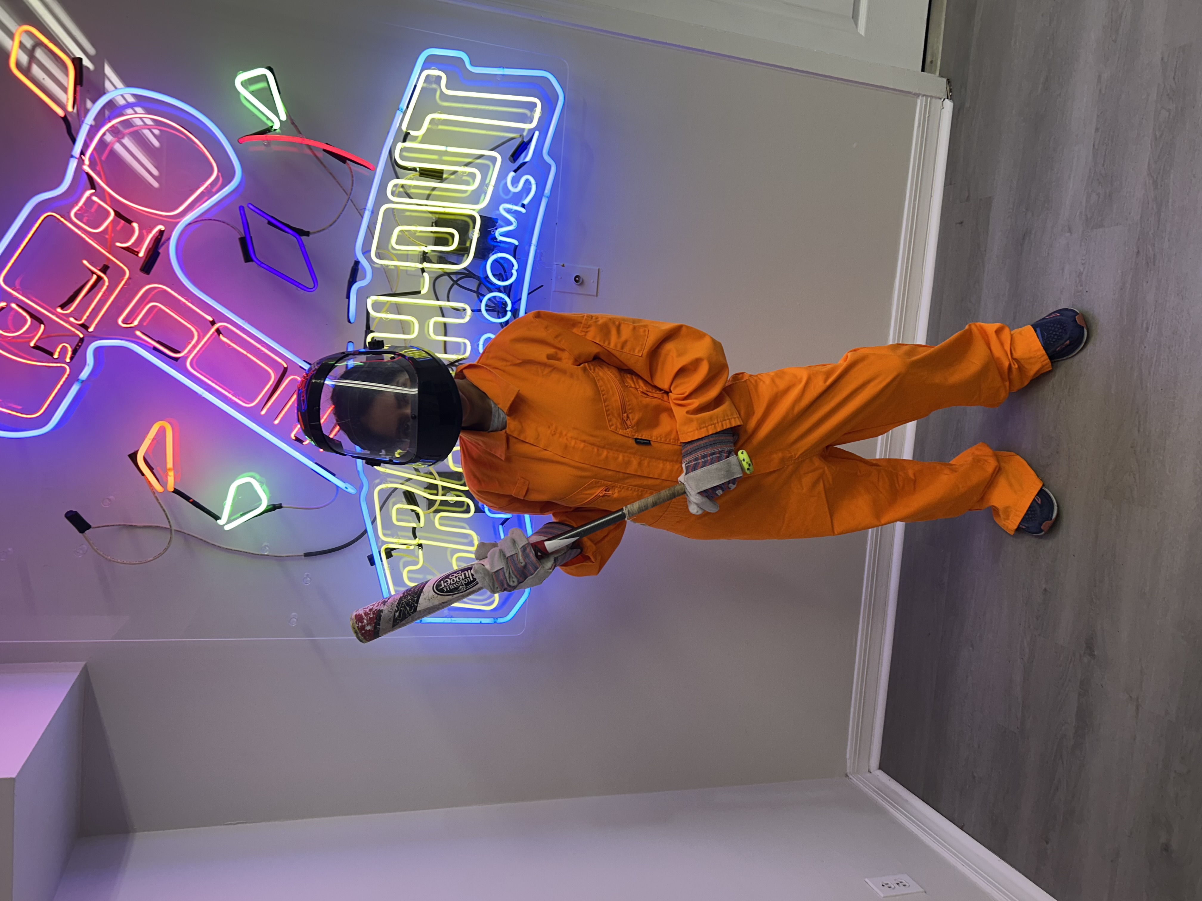 Crash Out customer in orange jumpsuit posing with baseball bat in front of vibrant Crash Out Rage Rooms neon sign