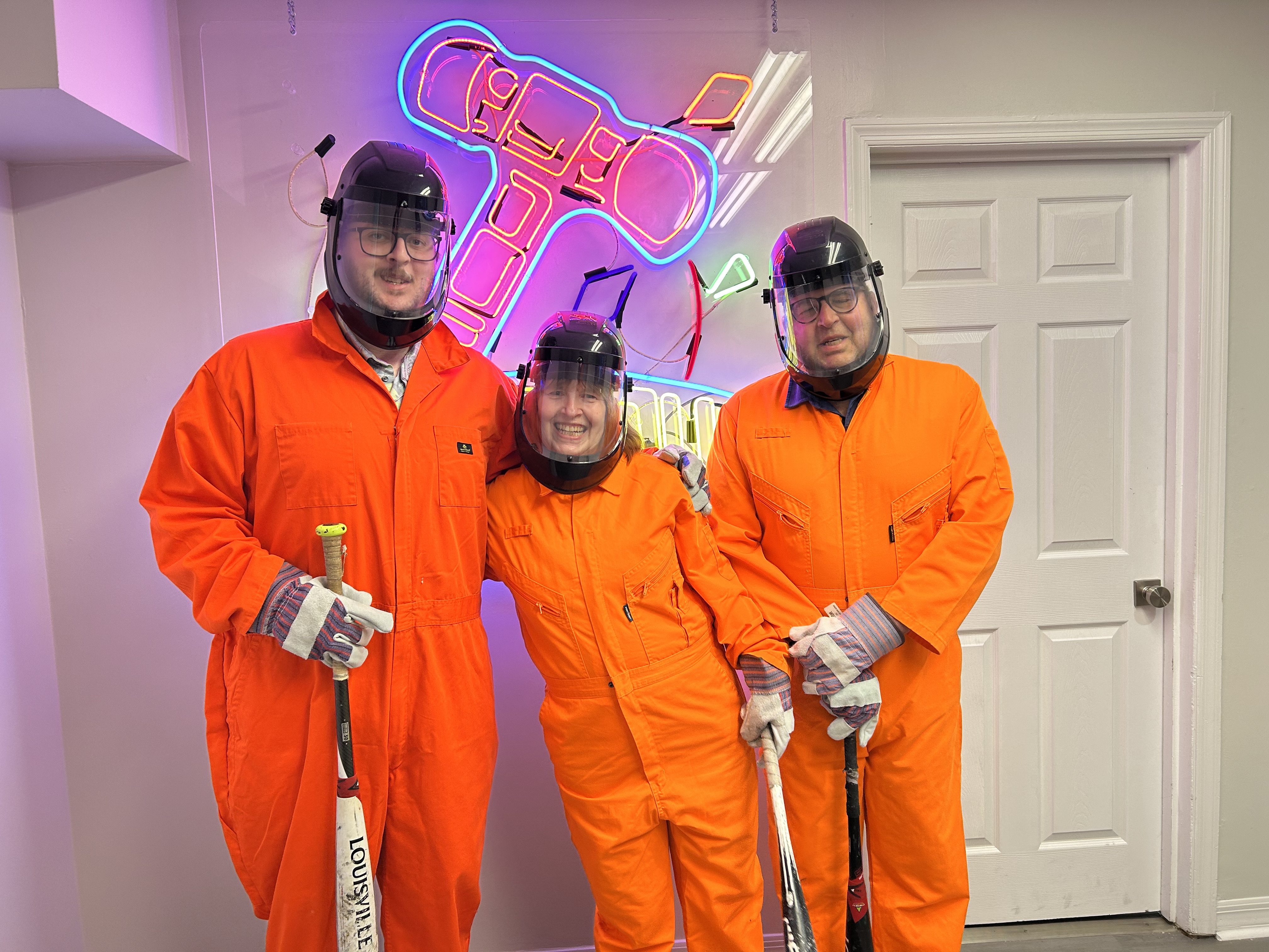 Three Crash Out guests in orange jumpsuits and face shields smiling and ready for their smash room session